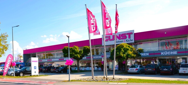 Ansicht der Möbelhauses GUNST von der Straßenseite aus. Bkauer Himmel, Parkplätze und 5 wehende Fahnen mit Logoschriftzug.
