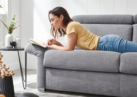 Möbel GUNST - Comfortmaster: Eine Frau liegt bäuchlings auf einem grauen, eingeklappten Schlafsofa und liest ein Buch.