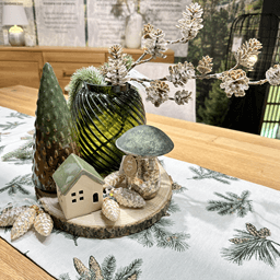 Tischdekoration aus Naturholz und Accessoires in Waldgrün: Baumscheibe als Unterlage, darauf eine grüne Vase mit Kiefernzweig, grüne Kerze sowie Holzdekoelemente (Baum, Pilz) und silbernen Glas-Zapfen.