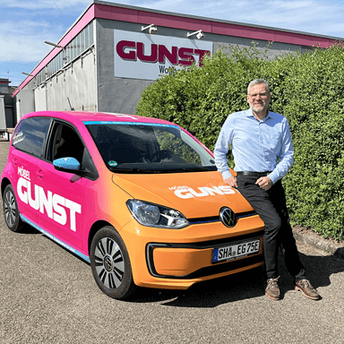 Geschäftsführer Tom Gunst hockt auf Motorhaube des Firmenfahrtzeugs, ein VW E-up im CD des Möbelhauses.  