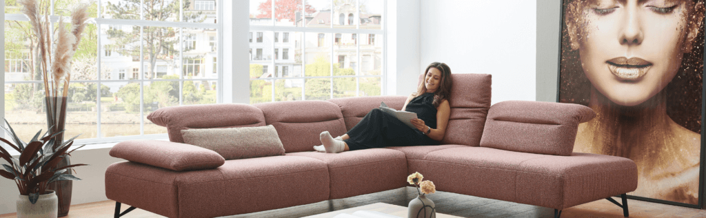 Eine Frau lehnt in der Sitzecke eines rostroten Ecksofas an einer hochgeklappten Rückenlehne und liest.