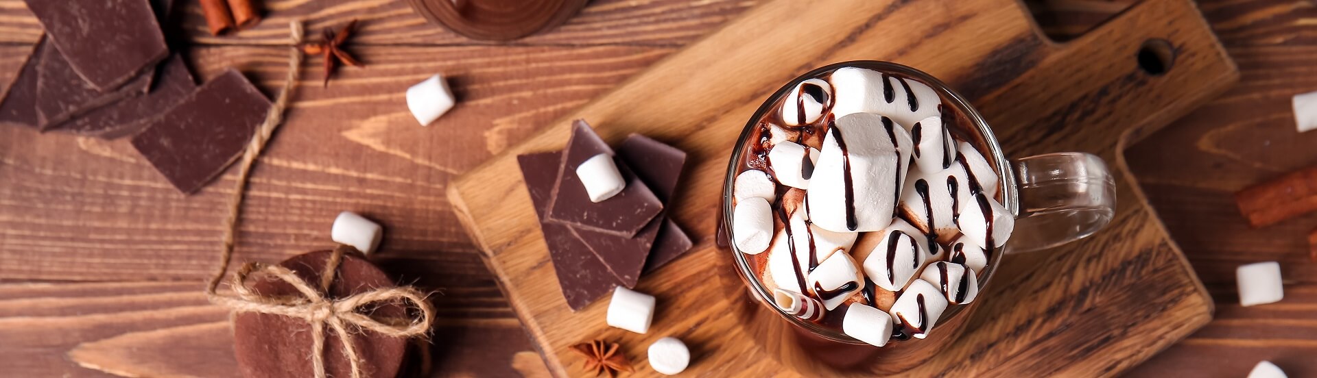 Sicht von Oben auf ein Glas heiße Schokolade, die mit vielen Marshmellows geschmückt ist und auf einem HolzBrett serviert wird.