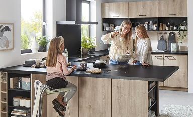 Eine Frau steht mit zwei Mädchen in einer offenen Küche und backt Pfannkuchen