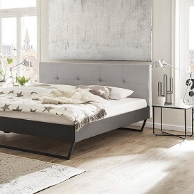 Schlicht-elegantes Boxspsringbett mit filigranen Metallfüßen, gepolstertem Kopfteil in hellgrau vor einer Wand mit Strukturtapete. Daneben ein schlichten Besitelltisch, der als Nachtkonsoe dient.