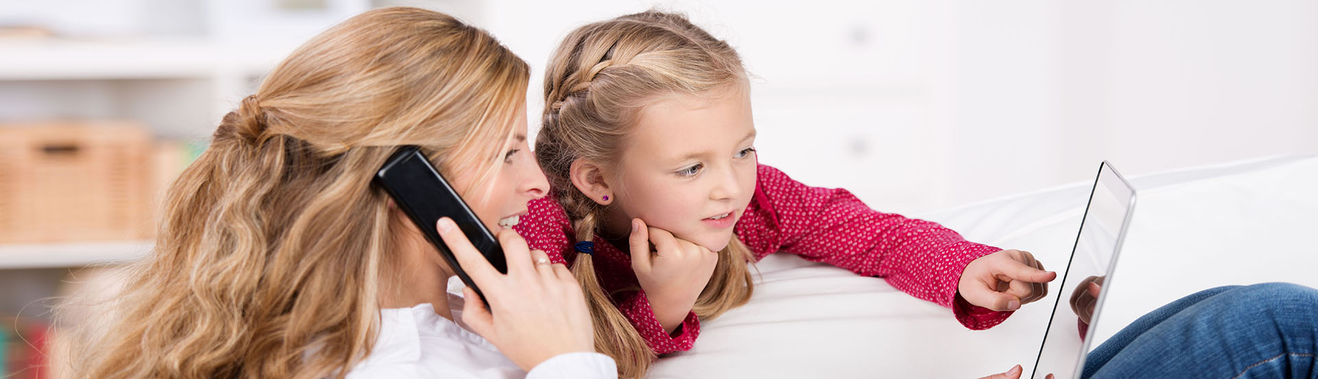 Mutter und Tochter sietzen im Sofa und schauen gemeinsam in eine Tablet. Die Mutter gibt eine telefonbestellung auf. 